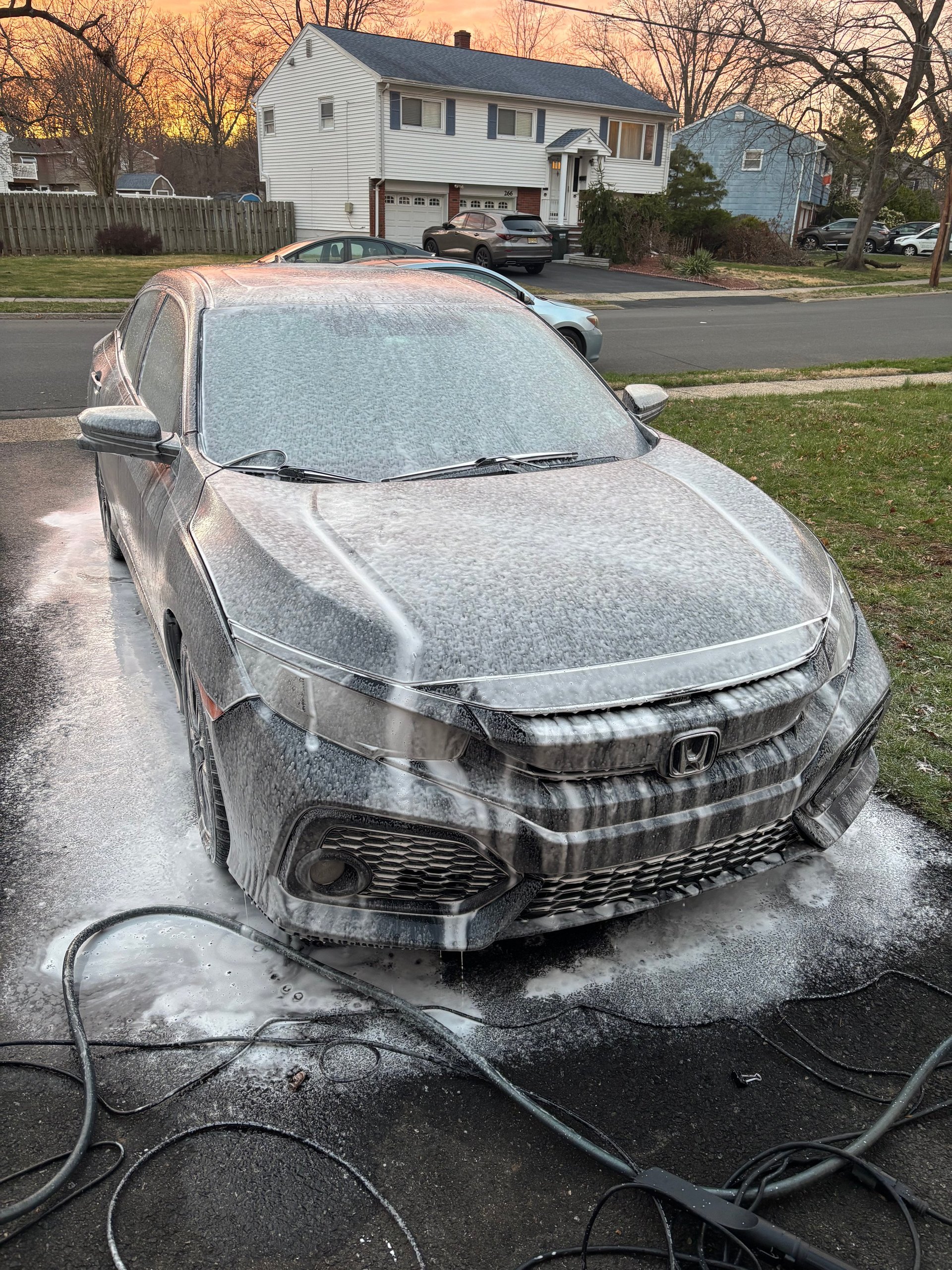 Silver Honda car being washed with soap and water in a residential driveway during golden hour